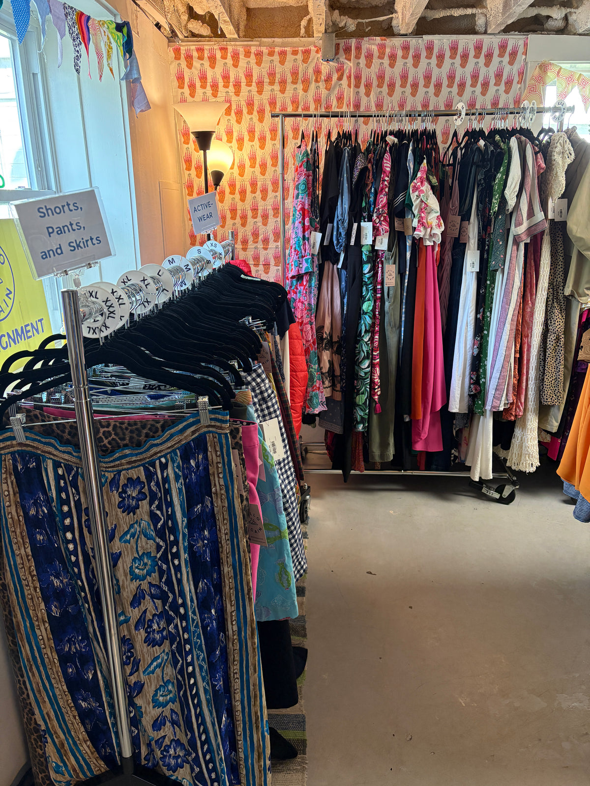 A boutique clothing store interior showing racks of colorful garments, patterned textiles on a table, and a sign that reads 'Shirts, pants, and skirts'.