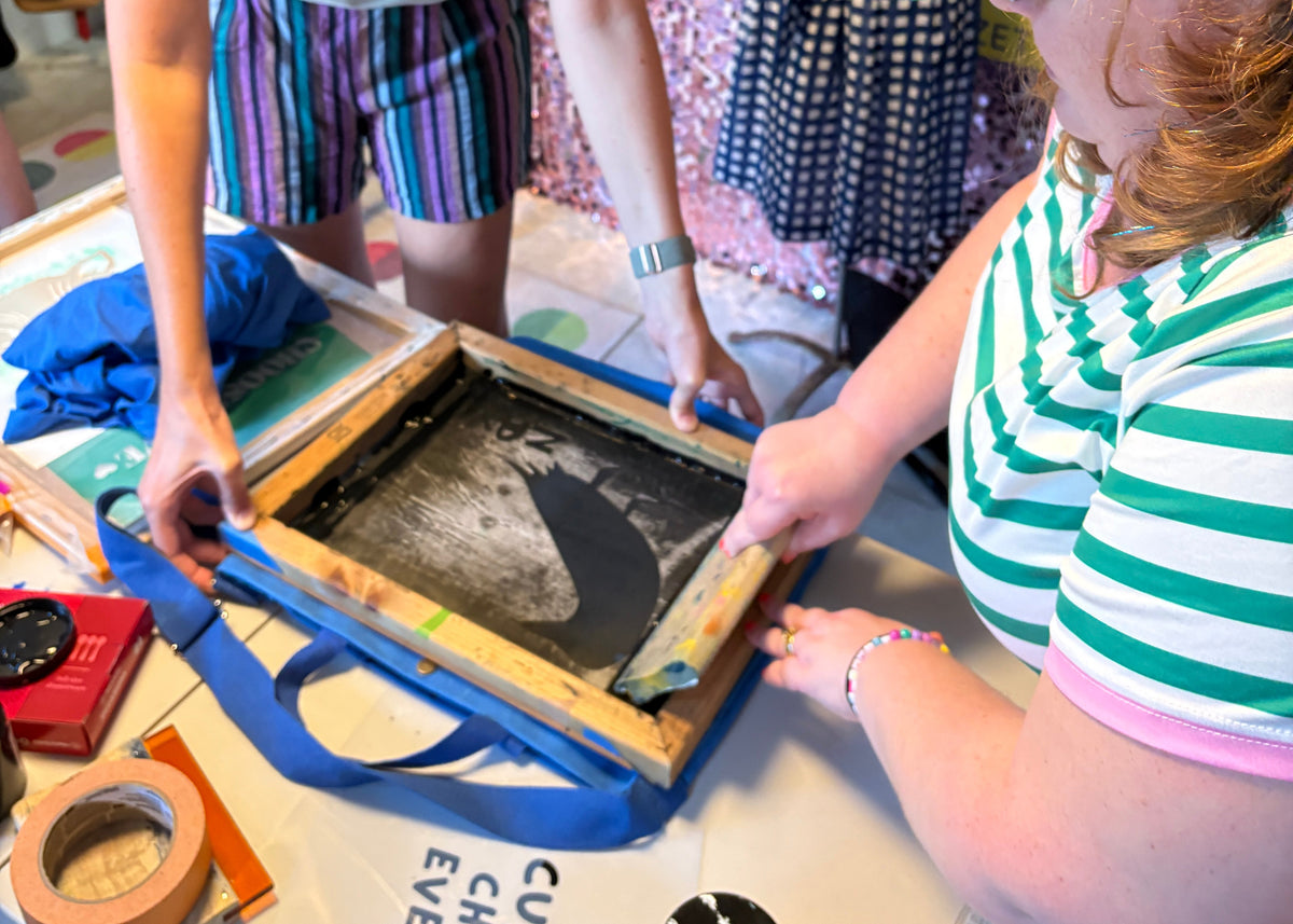 Two women collaborating at a table in a craft or retail shop, using a wooden screen printing frame with blue ink and fabric. One woman wears a green and white striped shirt and a green baseball cap, while the other wears a light gray t-shirt and colorful striped shorts.